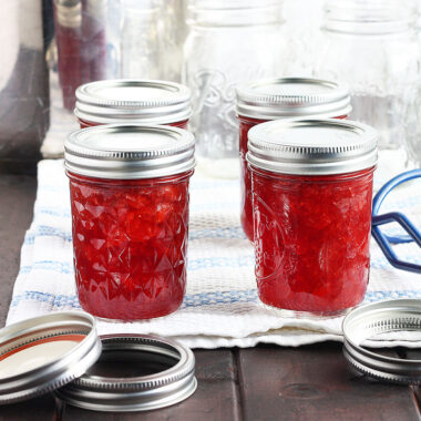 four jars of homemade jam cooling on a towel with empty mason jars and canning rings in the background.