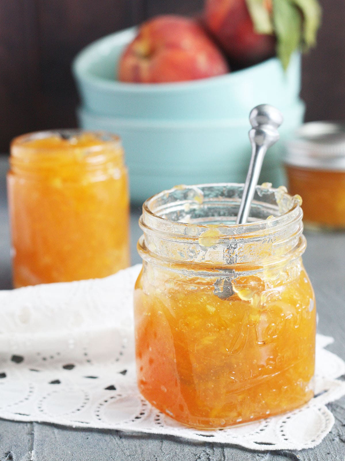 Two jars of homemade peach preserves with a small bowl of peaches in the background.