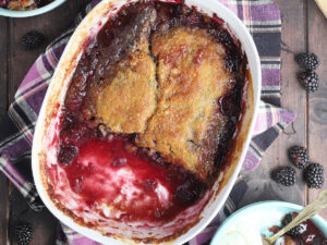 White serving dish of baked blackberry cobbler with two servings removed. Fresh blackberries are sprinkled around the sides.