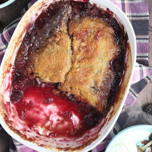 White serving dish of baked blackberry cobbler with two servings removed. Fresh blackberries are sprinkled around the sides.