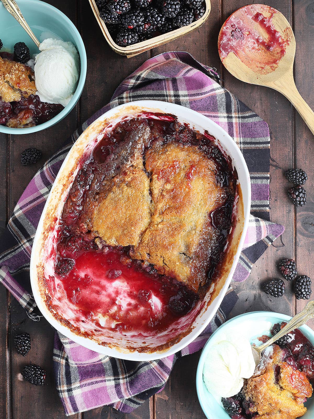 White serving dish of baked blackberry cobbler with two servings removed. Two blue bowls with cobbler and ice cream and fresh blackberries are sprinkled around the sides.