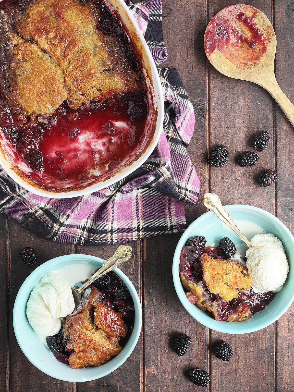 two blue bowls of blackberry cobbler with a scoop of vanilla ice cream. Serving dish of cobbler, a wooden serving spoon and fresh blackberries on the side