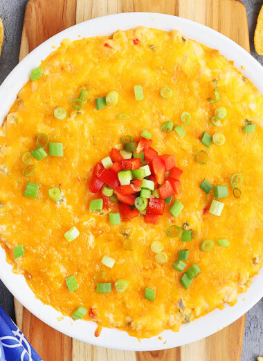 hot corn dip with cream cheese in a white pie plate garnished with green onions and red and green diced bell peppers. The plate rests on a wooden cutting board and corn chips are sprinkled to the side