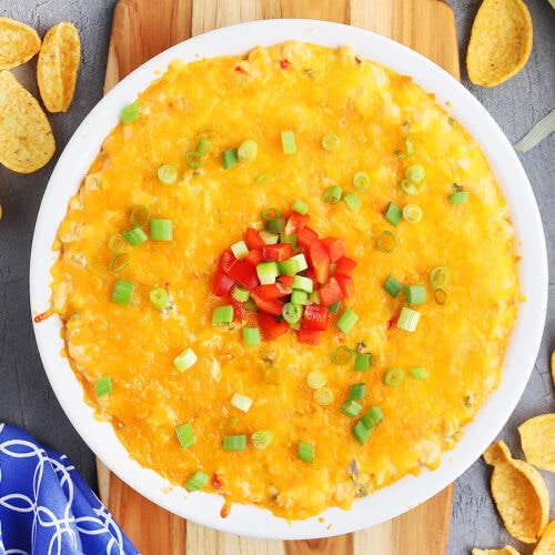 hot corn dip with cream cheese in a white pie plate garnished with green onions and red and green diced bell peppers. The plate rests on a wooden cutting board and corn chips are sprinkled to the side