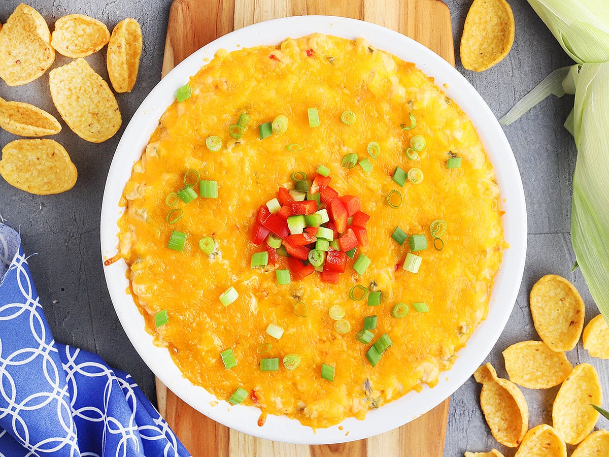 hot corn dip with cream cheese in a white pie plate garnished with green onions and red and green diced bell peppers. The plate rests on a wooden cutting board and corn chips are sprinkled to the side