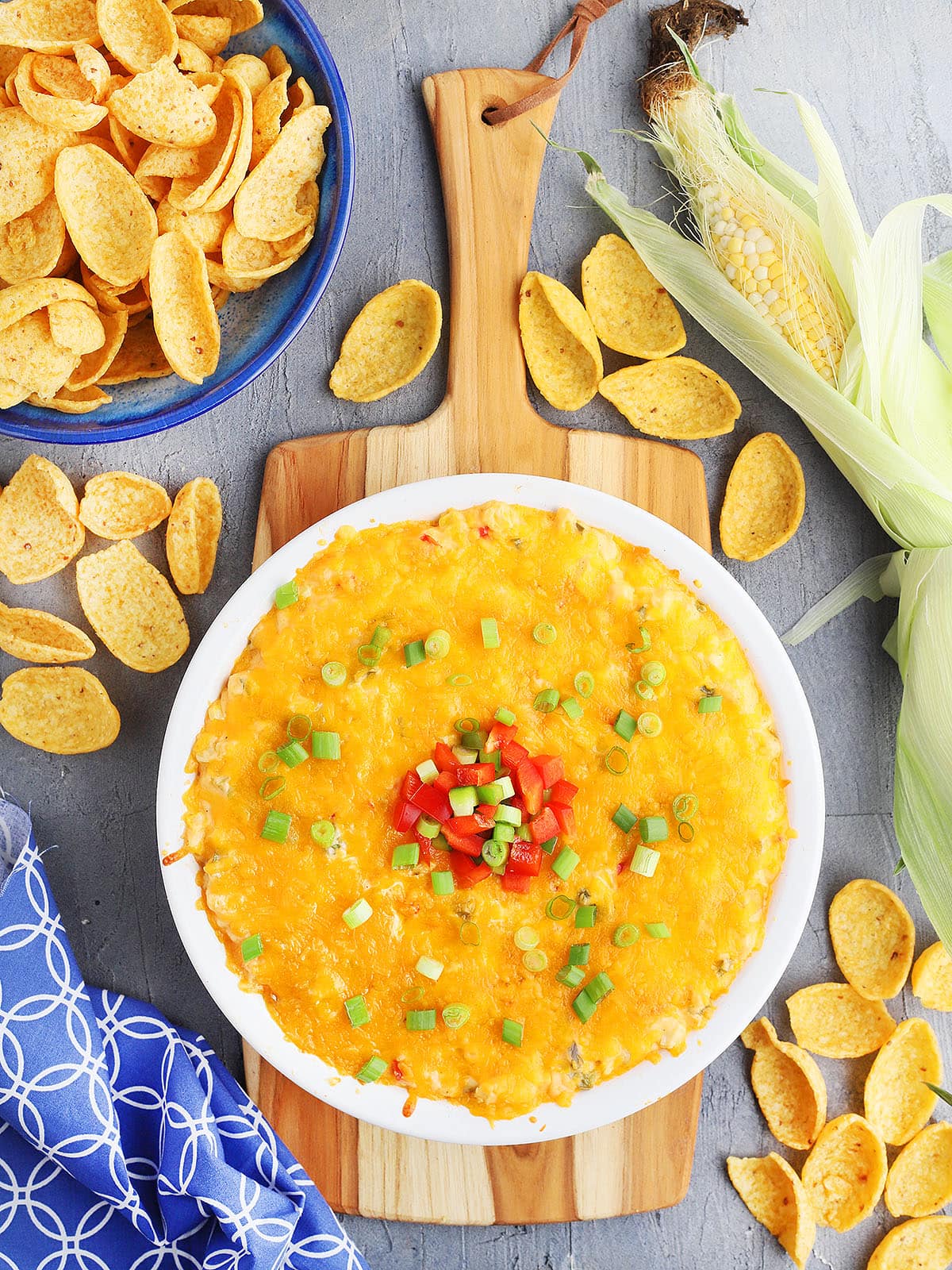 hot corn dip with cream cheese in a white pie plate garnished with green onions and red and green diced bell peppers. The plate rests on a wooden cutting board and a blue of corn chips sits to the side. Loose corn chips are sprinkled around.