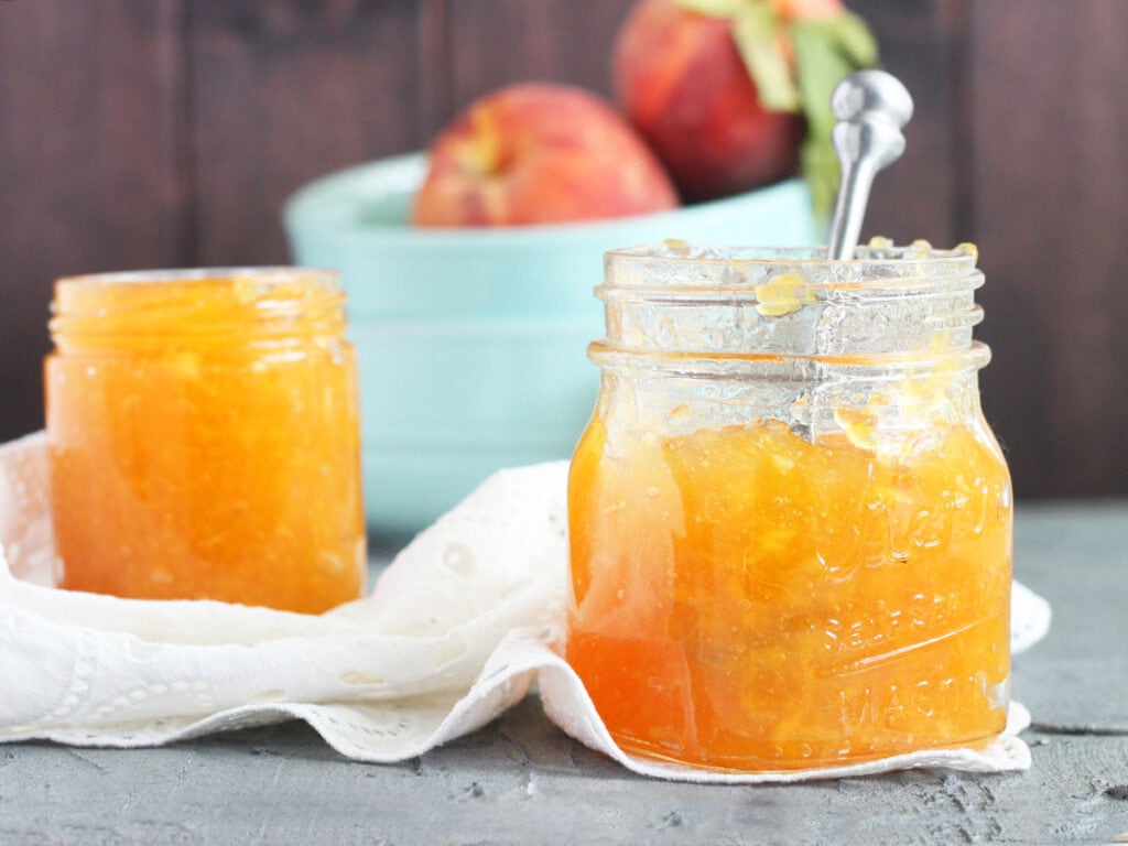 two jars of homemade peach preserves with a small bowl of peaches in the background.