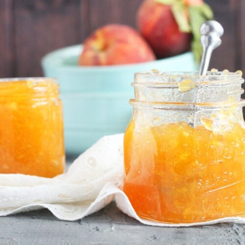 two jars of homemade peach preserves with a small bowl of peaches in the background.