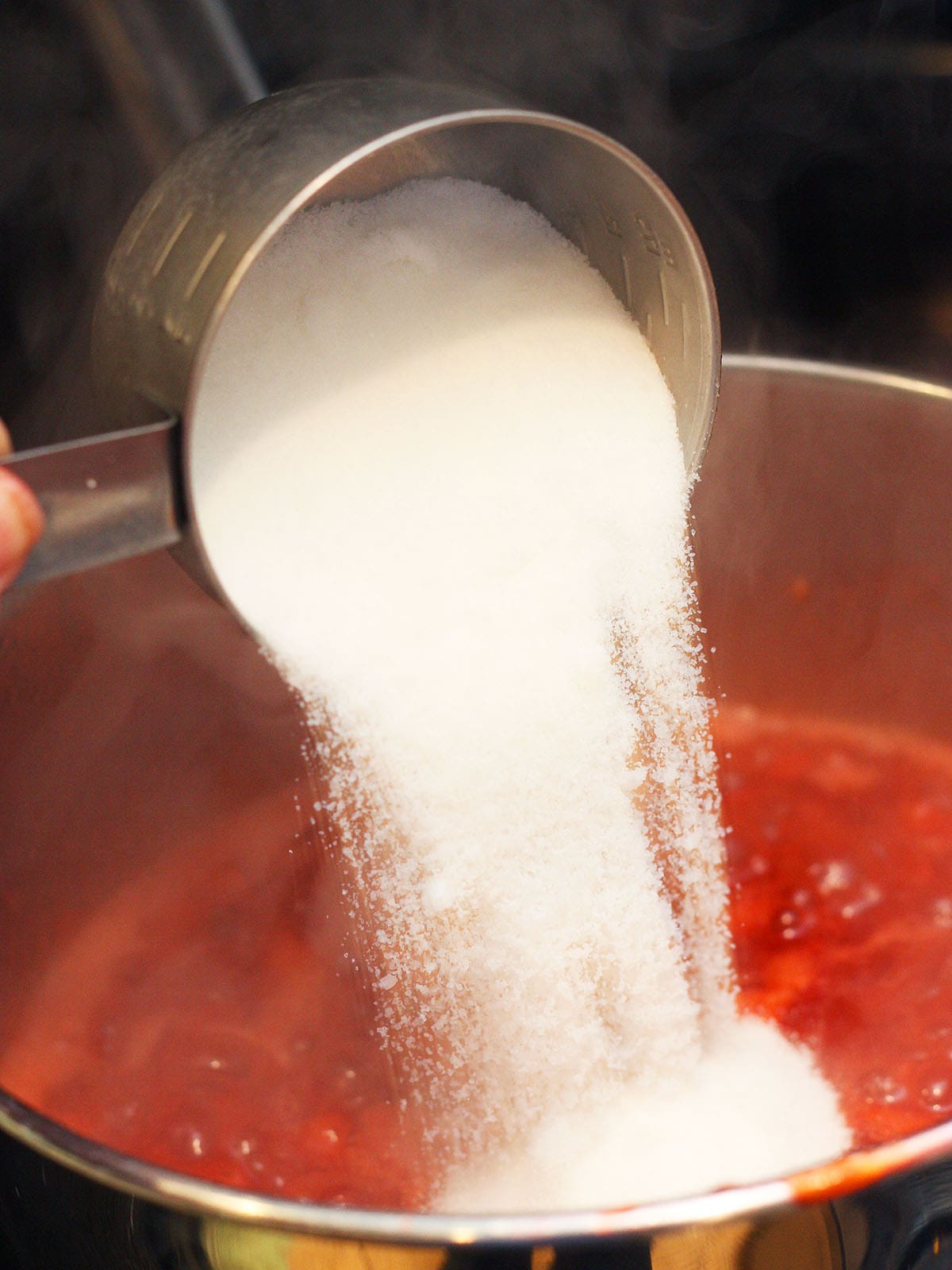 Metal measuring cup pouring white sugar into a pot of strawberry preserves.
