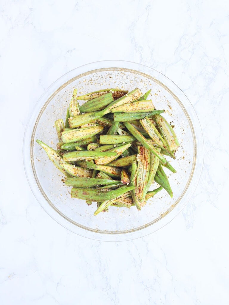 Sliced okra tossed with the seasoning mixture in a glass mixing bowl.