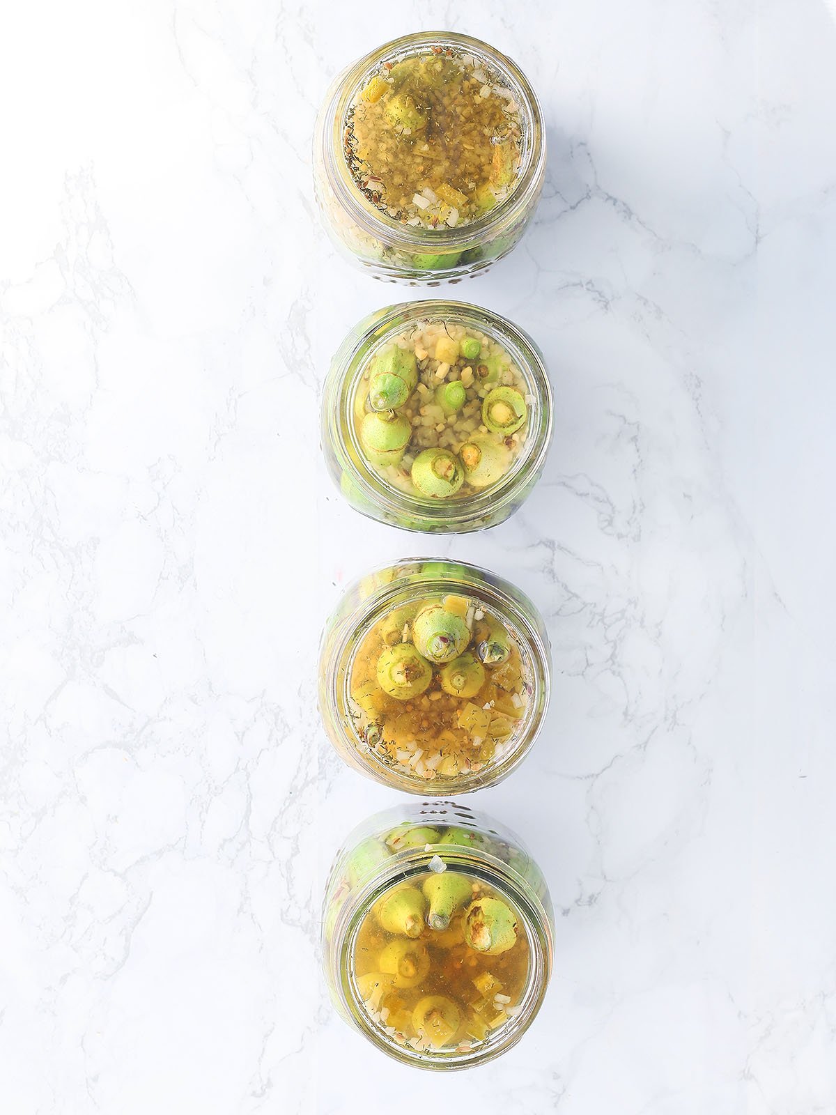 Four open jars of pickled okra lined up on a marble background.