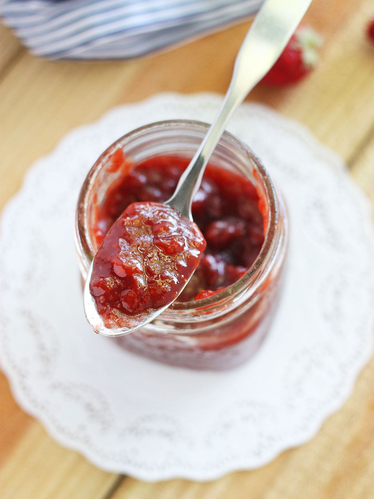 Close up shot of a glass jar of strawberry fig preserves on a white doily with a metal spoon of preserves resting on the top.