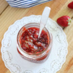 Glass jar of strawberry fig preserves on a white doily with a metal spoon sticking out.