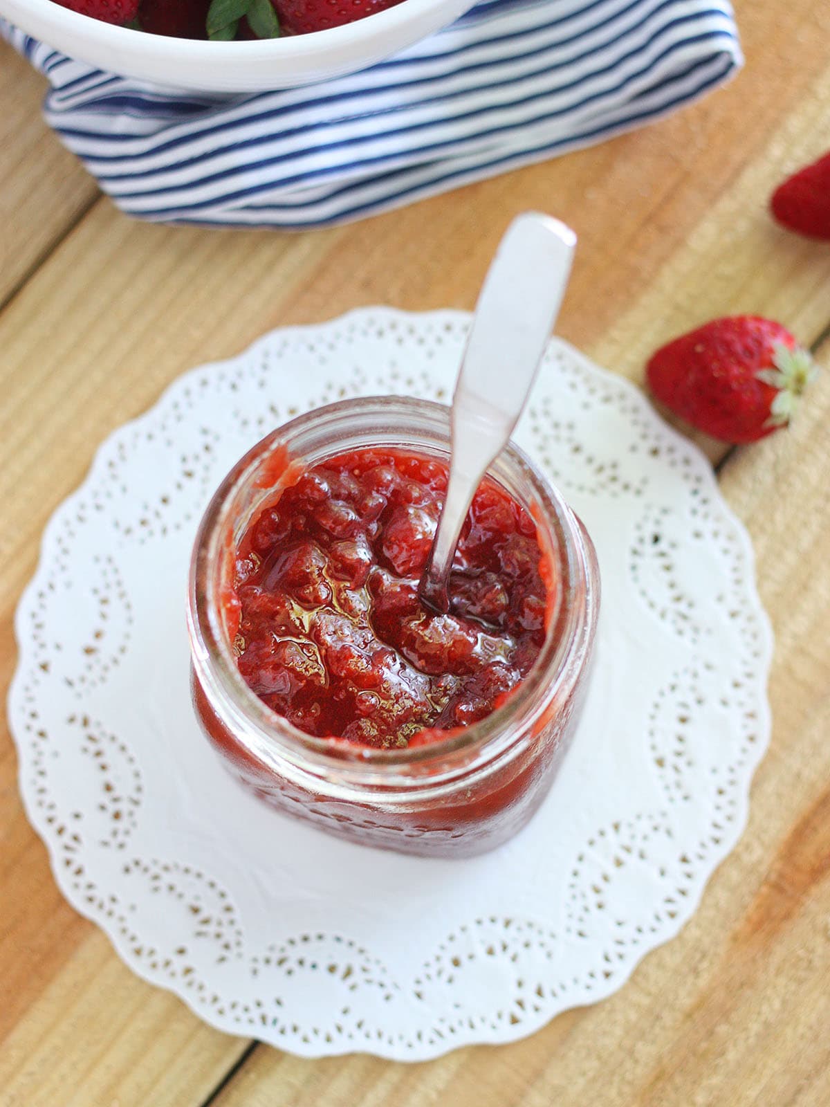 Glass jar of strawberry fig preserves on a white doily with a metal spoon sticking out.