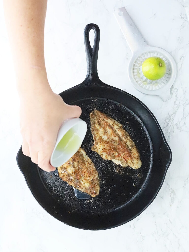 Hand pouring lemon juice over two catfish fillets in a cast iron skillet.