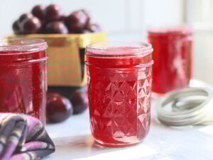 Three jars of muscadine jelly on a white counter top with a basket of fresh muscadines in the background.
