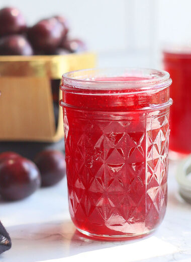 Three jars of muscadine jelly on a white counter top with a basket of fresh muscadines in the background.