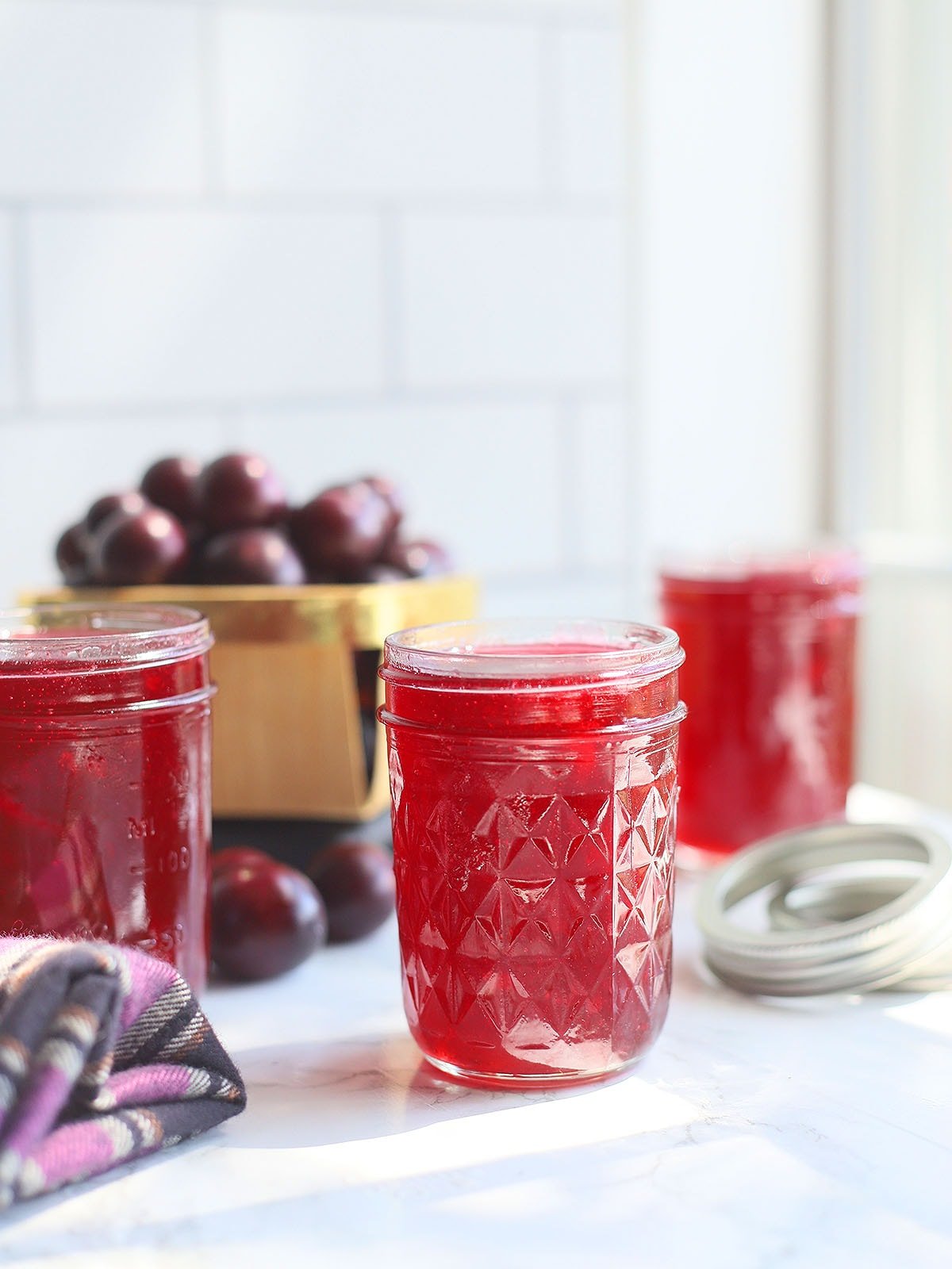 Three jars of muscadine jelly on a white counter top with a basket of fresh muscadines in the background.