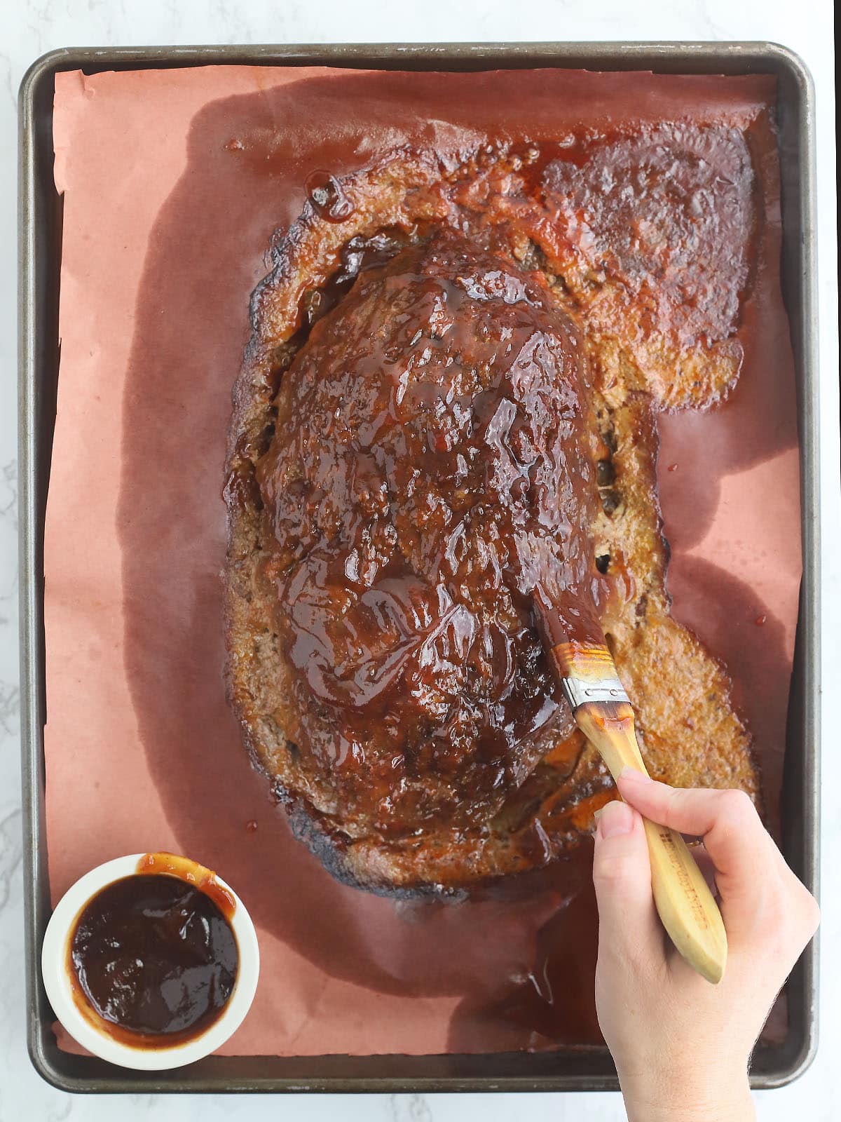 Hand brushing the top of a meatloaf with barbecue sauce.