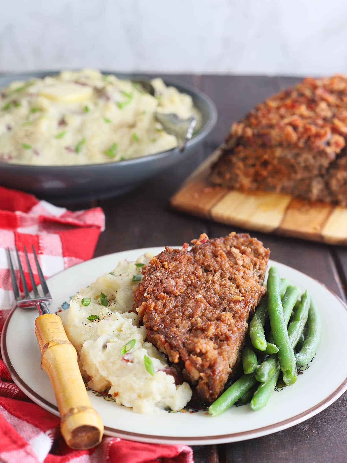 Slice of BBQ meatloaf on a plate with mashed potatoes and green beans.