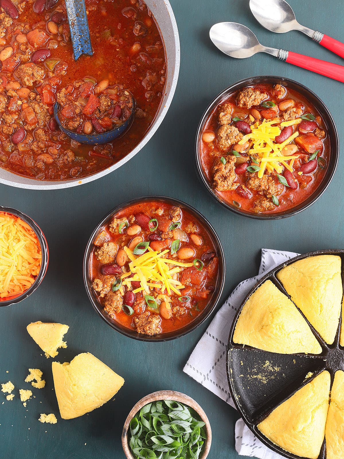 Two bowls of Cowboy Chili topped with shredded cheddar cheese with a pot of chili, cornbread and condiments to the side.