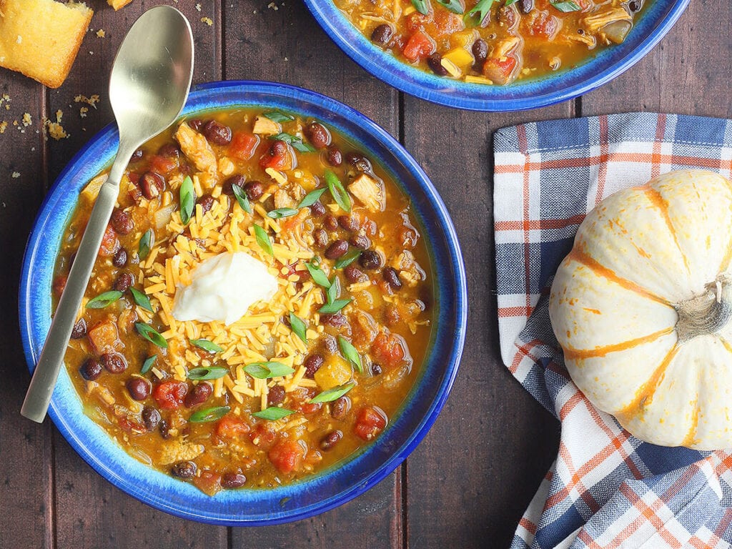 Two bowls of turkey pumpkin black bean chili garnished with shredded cheddar and sour cream.