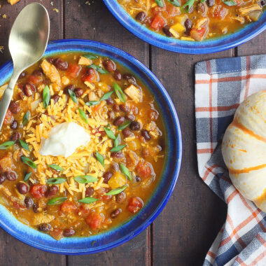 Two bowls of turkey pumpkin black bean chili garnished with shredded cheddar and sour cream.