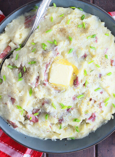 Garlic red skinned mashed potatoes topped with a pat of butter in a large serving bowl. A plate with a serving of potatoes and slice of meatloaf to the side.