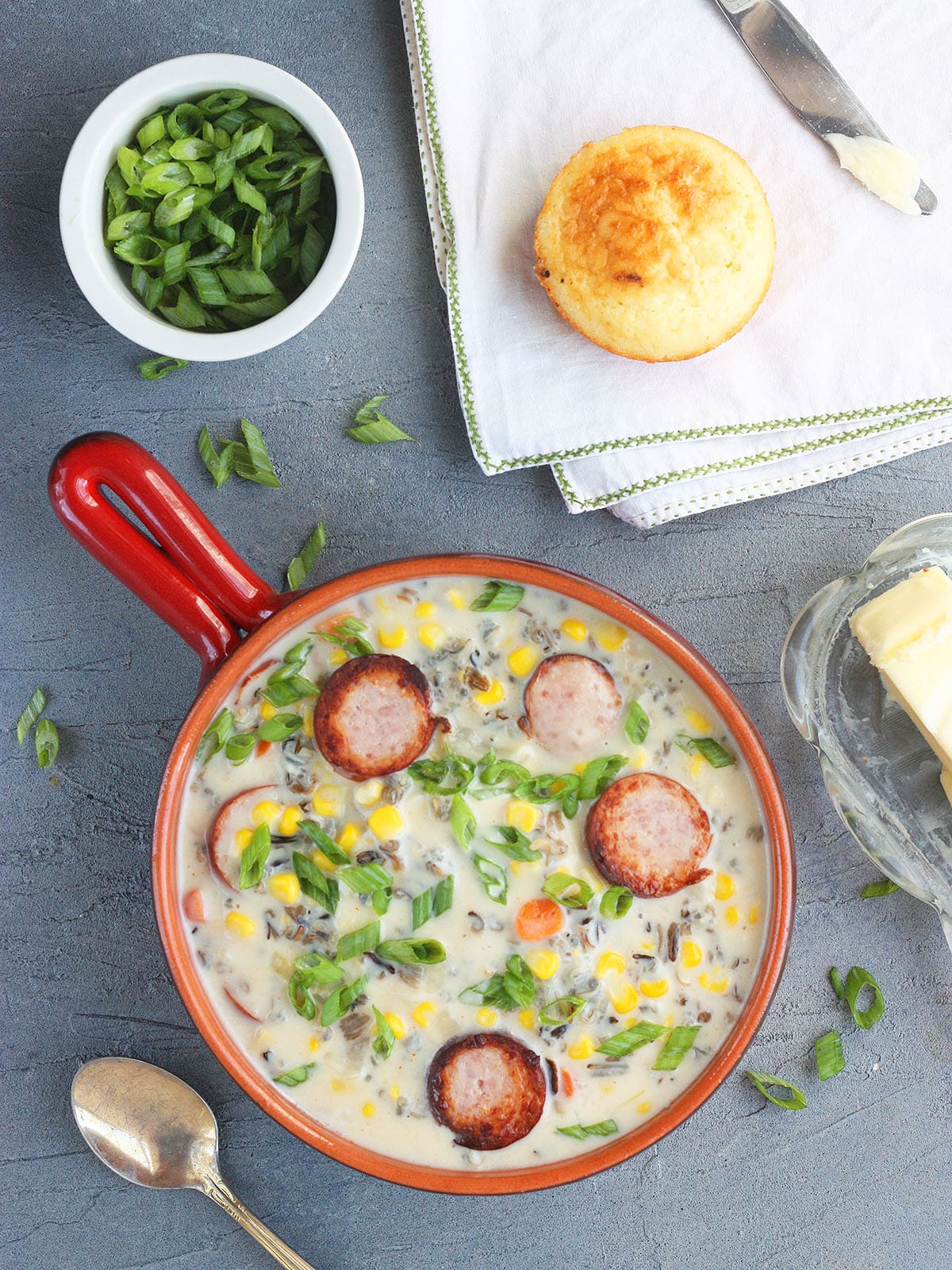 Serving of smoked sausage corn chowder garnished with green onions. A bowl of sliced green onions and a corn muffin are seen to the side.