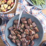 Serving dish of air fryer steak tips with a bowl of roasted potatoes, glass of wine and a platter of asparagus on the side.