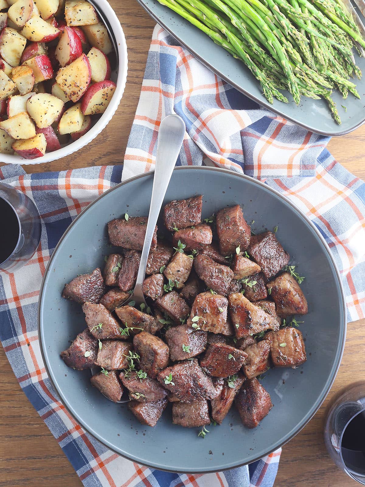 Serving dish of air fryer steak tips with a bowl of roasted potatoes, glass of wine and a platter of asparagus on the side.