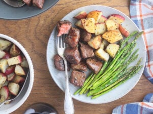Fork stuck in a medium rare steak tip resting on a plate with steak tips, roasted potatoes and asparagus.