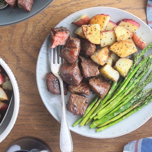 Fork stuck in a medium rare steak tip resting on a plate with steak tips, roasted potatoes and asparagus.