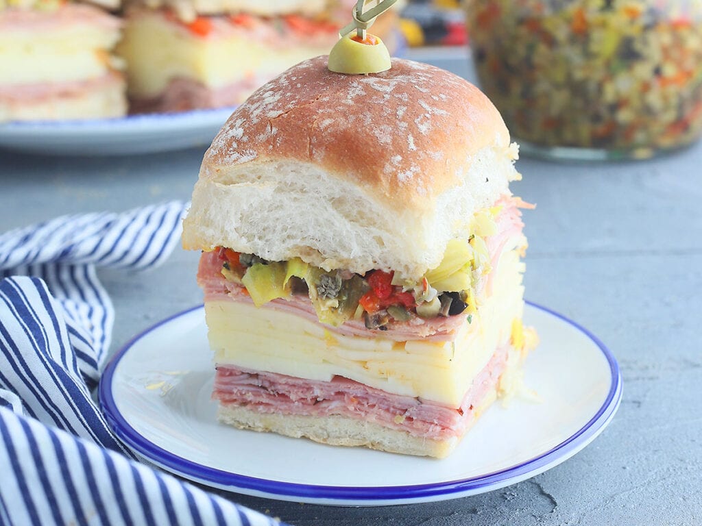 Single mini muffuletta sandwich on a small plate with additional sandwiches and a jar of olive salad in the background.