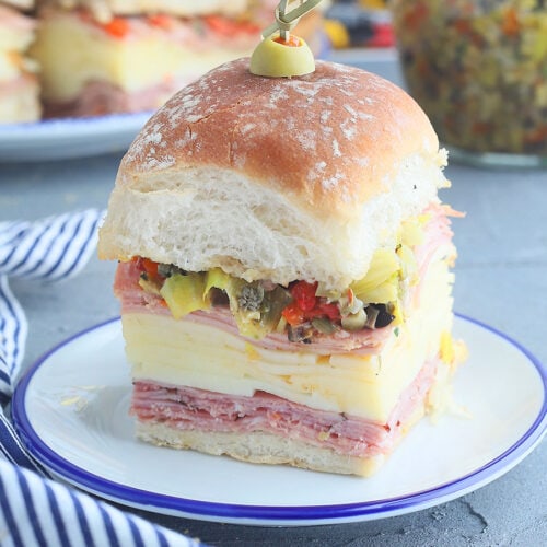 Single mini muffuletta sandwich on a small plate with additional sandwiches and a jar of olive salad in the background.
