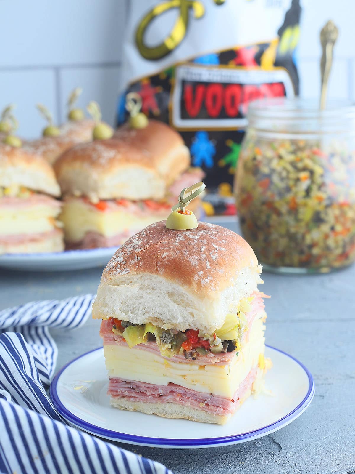 Single mini muffuletta sandwich on a small plate with additional sandwiches, a jar of olive salad and a bag of Zap's potato chips in the background.