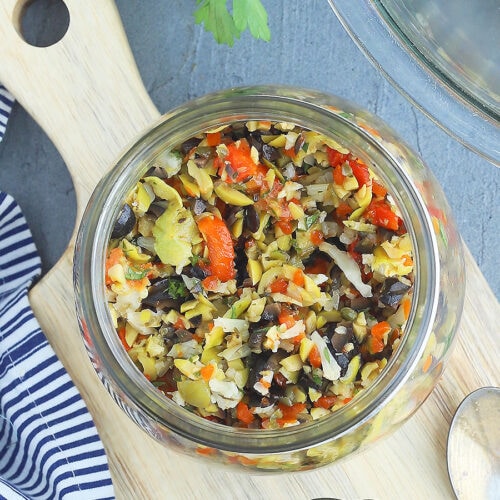 Looking down into an open jar of muffuletta olive salad setting on a wooden cutting board.