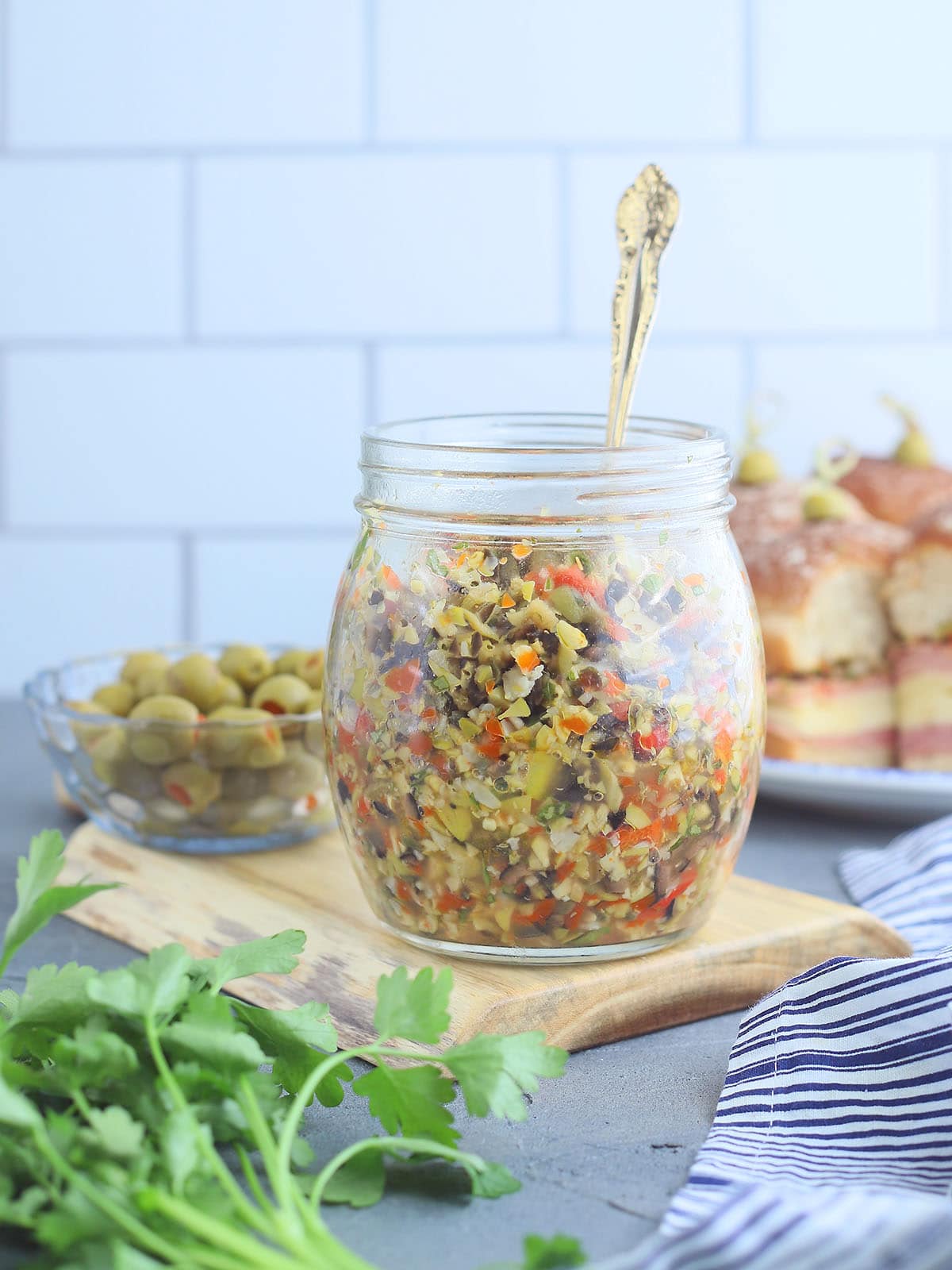 Glass jar of muffuletta olive salad with a serving spoon on a cutting board.