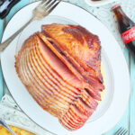 Sliced ham with Coca-Cola glaze on a white serving platter with side dishes and bottles of Coca Cola on the side.
