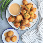 Platter of fried corn hushpuppies on a platter with dipping sauce and three hush puppies on a small plate on the side.