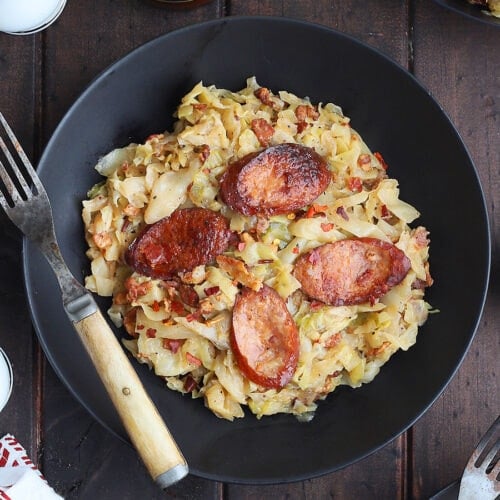 Bowl of Southern fried cabbage and smoked sausage with bacon and a rustic fork with hot sauce and a basket of cornbread muffins on the side.