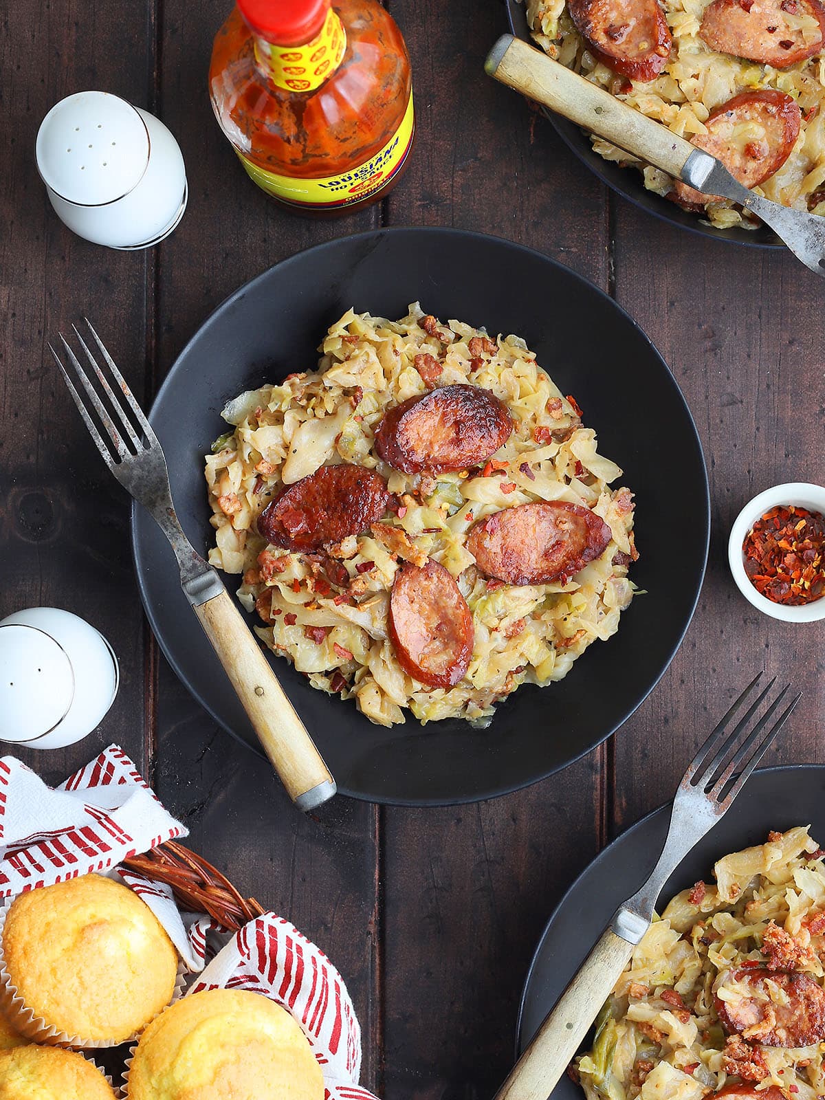 Bowl of Southern fried cabbage and smoked sausage with bacon and a rustic fork with hot sauce and a basket of cornbread muffins on the side.