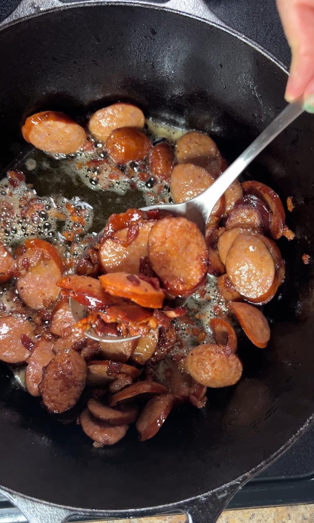 Using a slotted spoon to remove the fried smoked sausage and bacon from a Dutch oven.