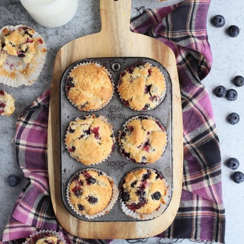 Buttermilk blueberry muffins in a muffin tin on a wooden cutting board with fresh blueberries and additional muffins on the side.