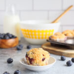 Single buttermilk blueberry muffin on a small dish with mixing bowls and baking ingredients in the background.