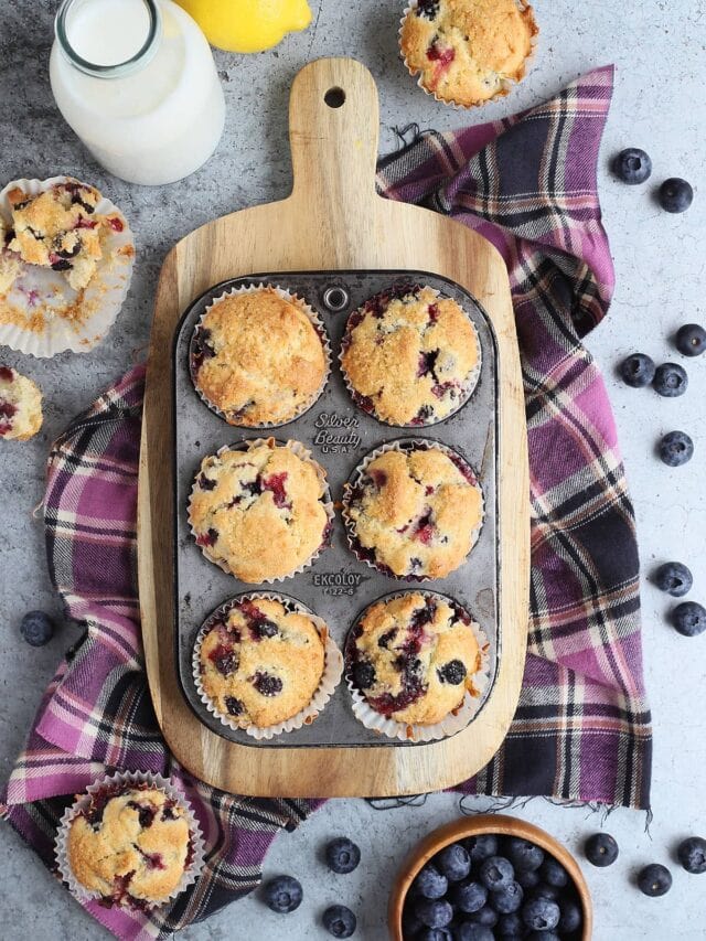 Buttermilk blueberry muffins in a muffin tin on a wooden cutting board with fresh blueberries and additional muffins on the side.