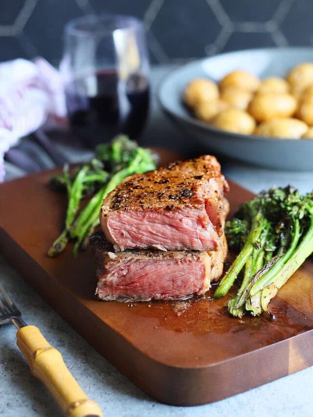 Pellet grilled steak cooked to rare cut in half on a wooden cutting board surrounded by roasted broccolini.