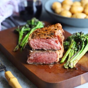 Pellet grilled steak cooked to rare cut in half on a wooden cutting board surrounded by roasted broccolini.