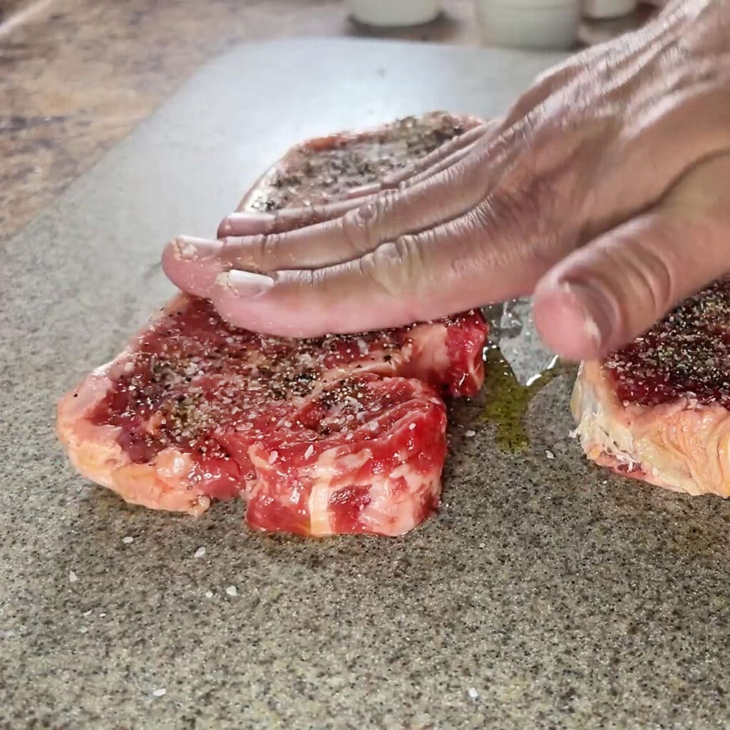 Patting seasonings into the surface of an uncooked New York strip steak.
