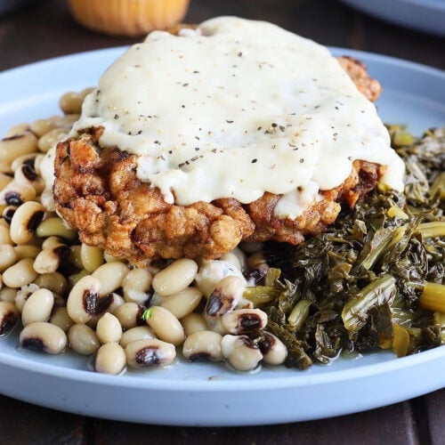 Chicken fried steak topped with country gravy on a plate with a pile of black-eyed peas and turnip greens.